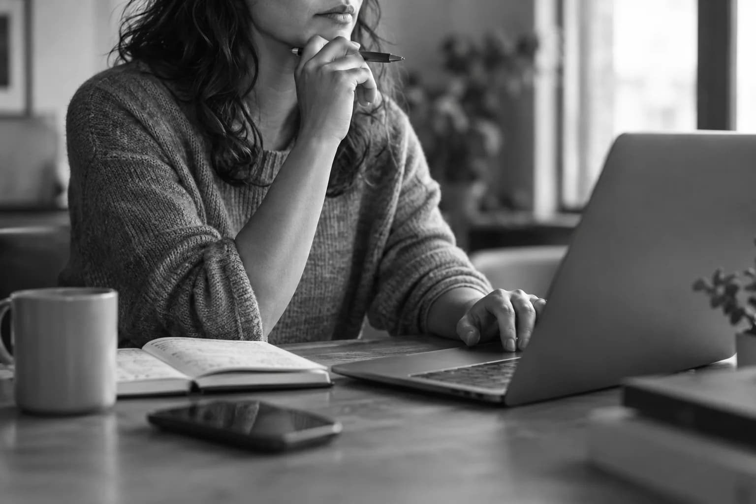 Monochrome photograph of a person reviewing a laptop and notebook by a window